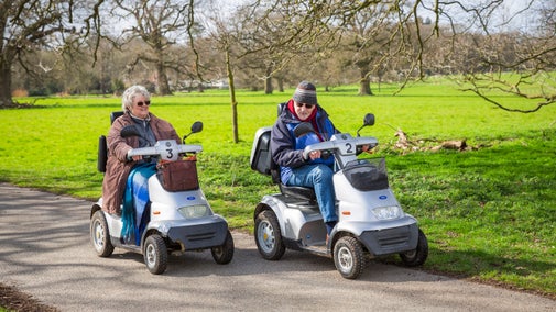 An elderly man and an elderly woman on two mobility vehicles parked side by side on a multi-use trail